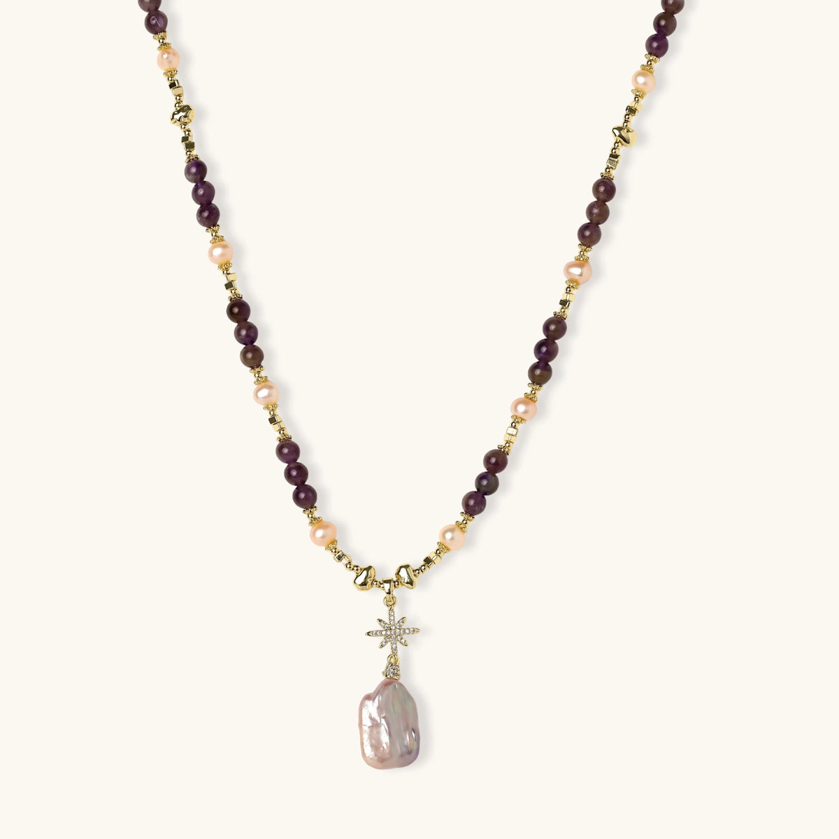 Gold necklace with iridescent baroque pearl pendant, purple amethyst beads, and crystal star charm on cream background